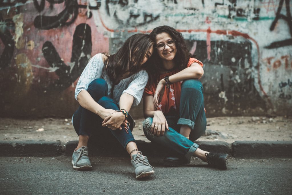 Zwei junge Frauen sitzen lachend auf einem Bürgersteig, die eine hat ihren Kopf an den Kopf der anderen gelehnt. Beide sehen sehr glücklich aus.