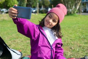 Mädchen mit lila Jacke und Mütze macht lächelnd ein Selfie mit einem Smartphone